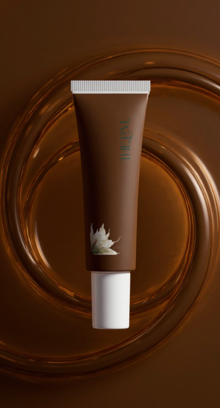 A brown cosmetic tube with a white cap, featuring a white lotus flower illustration and the brand name 'the p.m.' printed vertically, set against a glossy brown background with circular light reflections.