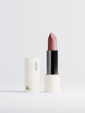 A lipstick bullet in a dusty mauve-pink shade stands upright next to its white cylindrical cap, which features the brand name 'THE P.M.' printed vertically. The lipstick has a glossy black center body and sits on a clean, light gray studio background.