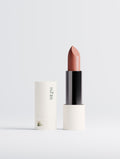 A lipstick bullet in a warm dusty rose shade stands upright next to its white cylindrical cap, which features the brand name 'THE P.M.' printed vertically. The lipstick has a black center body and is set against a clean, light gray studio background.
