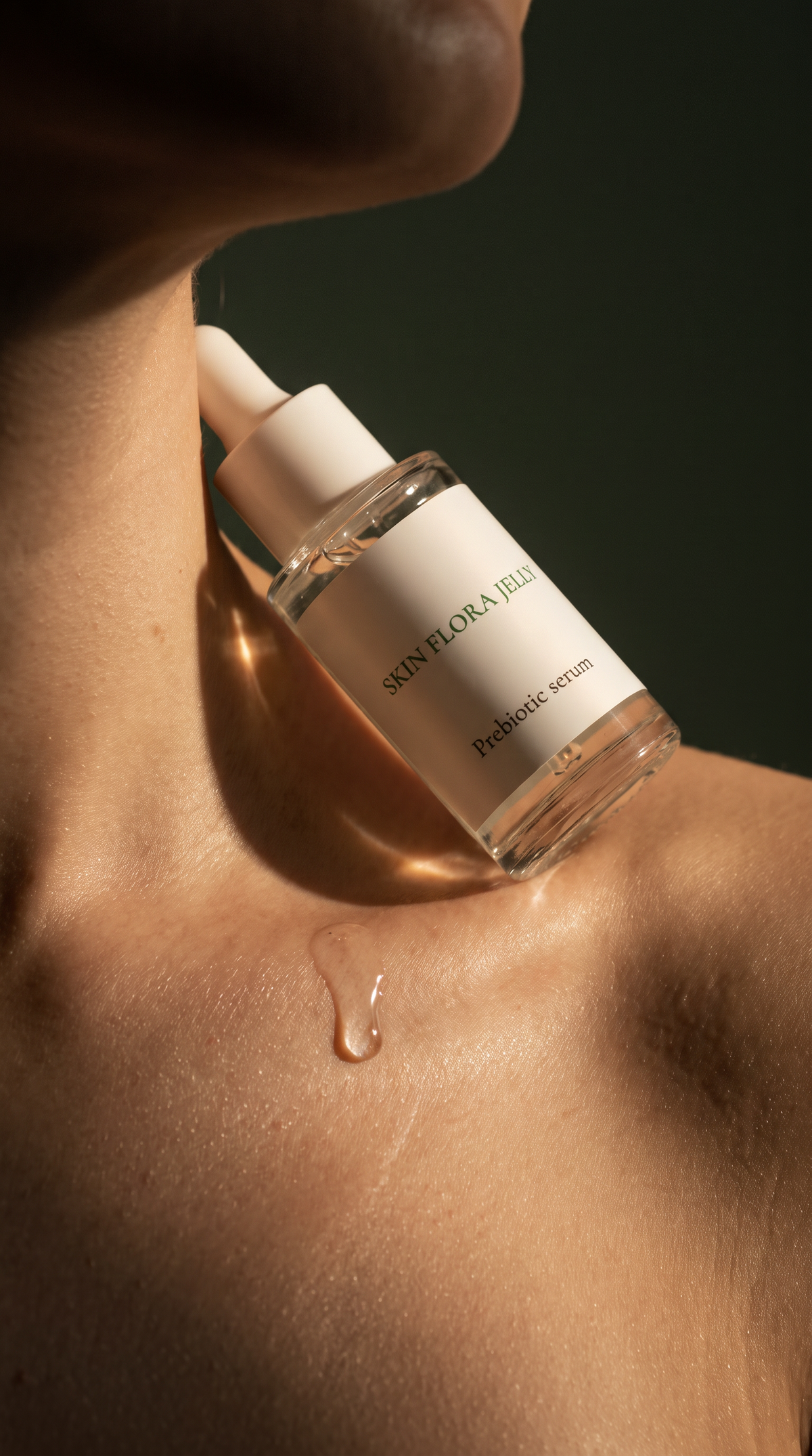A close-up of a person's neck and shoulder with a clear dropper bottle labeled 'SKIN FLORA JELLY Prebiotic serum' resting on the skin, with a glossy drop of serum visible against a dark background.